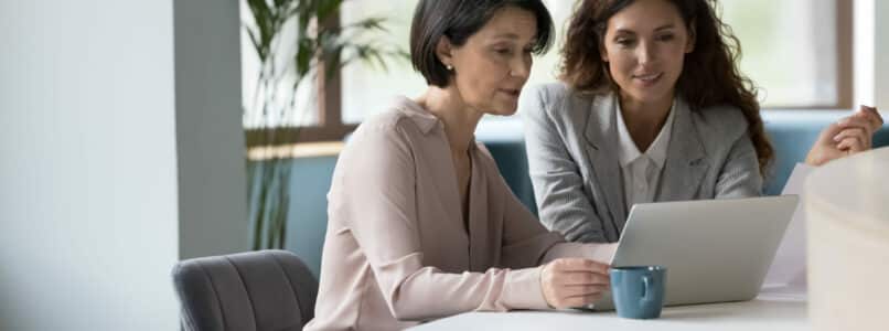 35s and 55s women colleagues working together use computer to learn AI, meet in office staring at laptop screen, AI training, HR Training, learn new software, younger Hispanic affiliate helps to older with program or app. Tech, business